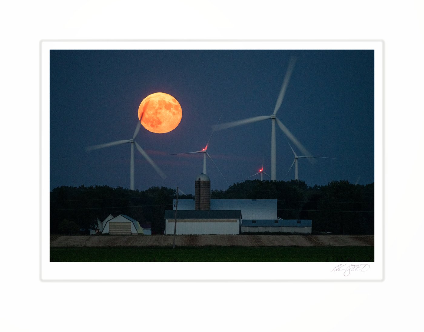 A supermoon rising orange behind wind turbines beyond a farm in the foreground. Near Caseville, Michigan -September 18, 2024